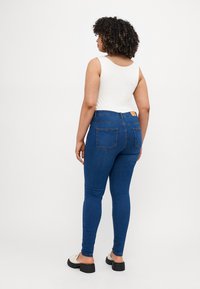 Skinny blue jeans with a high waistband, back pockets, and a smooth texture, paired with a white sleeveless top and light footwear.