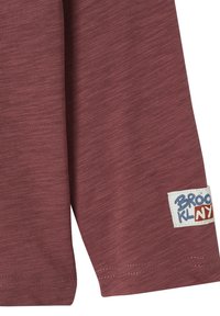 Langärmliges, burgunderfarbenes Baumwollhemd mit strukturiertem Oberflächen und einem Etikett, auf dem "BROOKLYN" in Blau und Rot auf weißem Hintergrund gedruckt ist.