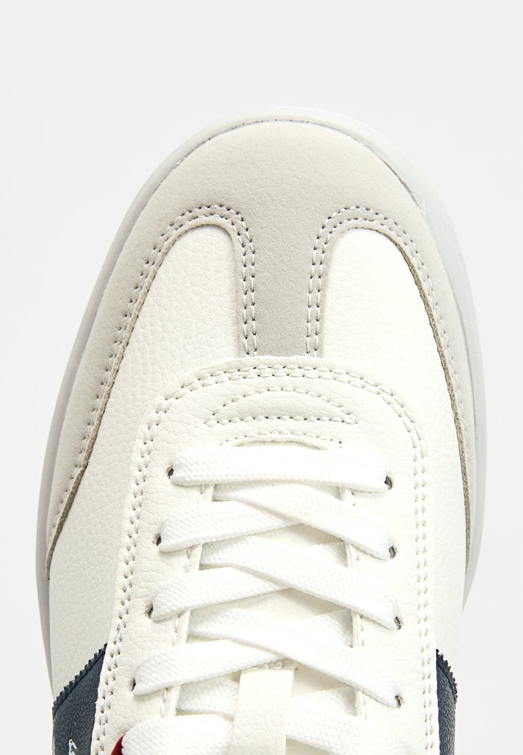 Zapatilla blanca de cuero con parte superior texturizada, diseño con cordones, acentos en gris y azul marino, y suela de goma. Presenta un detalle de costura en la puntera.