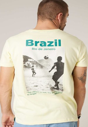 Mann trägt ein hellgelbes T-Shirt mit grünem Schriftzug "Brazil Rio de Janeiro" und einem schwarz-weißen Bild von Jungen, die am Strand Fußball spielen.
