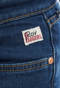 Tmavě modré džíny z denimu s bílou nášivkou s nápisem "Roy Rogers" v červeném a černém vyšívání, měděným knoflíkem a kontrastním oranžovým šitím.