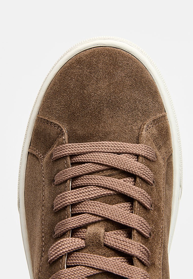 Braune Wildledersneaker mit rundem Zeh, kontrastierender weißer Gummisohle und flachen braunen Schnürsenkeln. Keine sichtbaren Logos oder Muster.