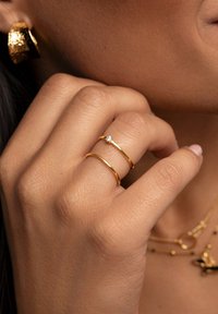 Anillos de oro con bandas suaves, uno de ellos presenta un pequeño diamante. Una mano muestra una piel texturizada junto a varias joyas de oro.