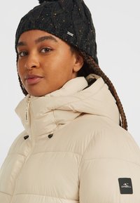 Beige Puffjacke mit hohem Kragen, schwarzen Hardware-Akzenten und einem Logopatch am Ärmel. Kombiniert mit einer schwarzen Strickmütze.