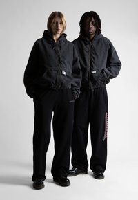 Juodos gobtuvinės striukės ir juodos kelnės, su elastinėmis rankovėmis. Striukės turi užtrauktuką ir priekinės kišenes; vienos kelnės turi grafinius akcentus.