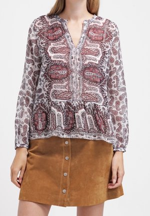 Vrouw draagt een blouse met lange mouwen en een patroon, met een gespleten halslijn, en een bruine suède rok met knopen aan de voorkant.