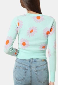 Maglione verde menta a maniche lunghe, con motivi a margherite rosa e arancioni, texture a coste e design aderente. Indossato con jeans.