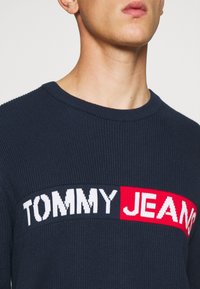 Granatowy prążkowany sweter z dzianiny z poziomym wzorem logo w bieli i czerwieni oraz wyraźnym napisem "TOMMY JEANS".