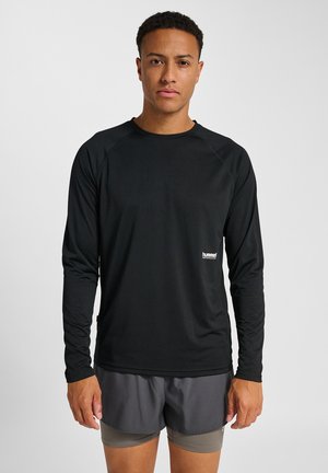 Man met een zwart, lang mouw prestatie shirt en grijze laagjes hardloopshorts, staand tegen een effen lichte achtergrond.