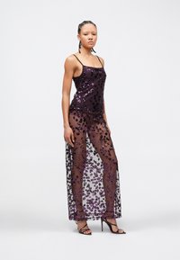 Femme debout portant un haut violet à bretelles fines en sequins et une jupe transparente assortie à motifs floraux, avec des sandales à talons noirs.