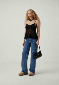Zwarte gebreide tanktop met een peplumontwerp, blauwe brede jeans, zwarte handtas met studs en lichtbruine klompen. Neutrale achtergrond.