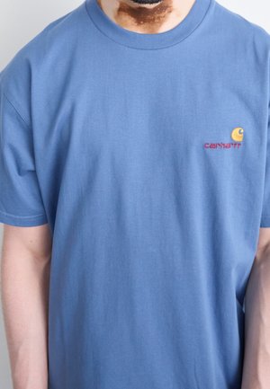 Man draagt een effen blauwe Carhartt T-shirt met korte mouwen, toont de bovenkant van de torso en een deel van de nek en armen.