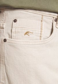 Jeans de mezclilla beige con un pequeño logo bordado en el bolsillo frontal, con detalles de costura sutil y acentos de botón metálico.