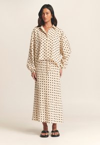 Cream-colored, long-sleeve shirt and matching skirt set with black polka dots; loose fit with buttons and drawstring accents. Black sandals.