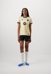 Camiseta de fútbol color amarillo claro con acentos en negro y azul, que presenta logotipos. Combinada con pantalones cortos negros, calcetas blancas y botas azules.