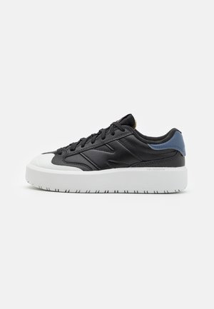 Schwarzer flacher Sneaker mit weißer dicker Sohle, abgerundetem weißem Zehenbereich, schwarzen Schnürsenkeln und blauem Fersenlaschen auf hellem einfarbigem Hintergrund.