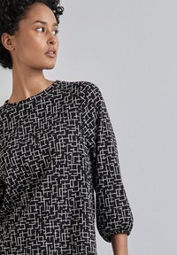 Schwarzes Kleid mit einem weißen geometrischen Muster, runder Ausschnitt und 3/4 Ärmeln; der Stoff wirkt weich und strukturiert. Verfügt über eine lässige Passform.