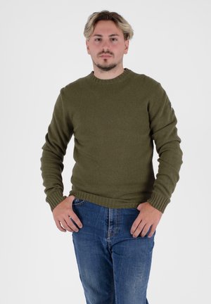 Olijfgroene gebreide trui met geribbelde textuur, ronde hals en lange mouwen; gecombineerd met blauwe denim jeans. Eenvoudig ontwerp en strakke pasvorm.
