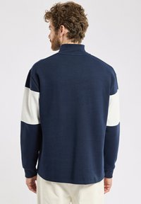 Sweatshirt bleu marine avec des panneaux latéraux blancs, col montant, poignets côtelés et tissu texturé. La vue arrière présente un design minimal.