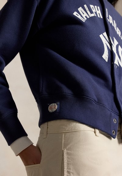 Πρόσωπο φοράει ένα σκούρο μπλε φούτερ Ralph Lauren με κονκάρδα Polo RL και μπεζ παντελόνια, χέρι στην τσέπη, σε απλό φόντο.