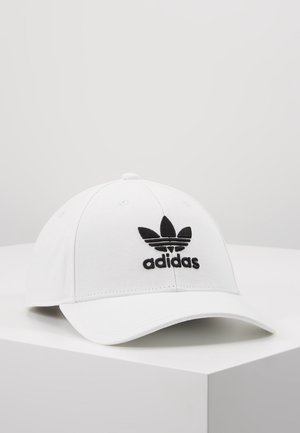 Biała czapka z daszkiem Adidas z czarnym logo trefoil i napisem, umieszczona na białej powierzchni na tle jasnoszarym.