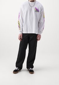 Witte longsleeve t-shirt met kleurrijke abstracte ontwerpen op de mouwen, gecombineerd met zwarte broek en zwarte sneakers.