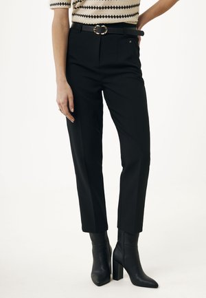 Schwarze maßgeschneiderte Hosen mit hoher Taille, ausgestattet mit Gürtelschlaufen und einer goldenen, runden Schnalle. Kombiniert mit enganliegenden schwarzen Stiefeletten.