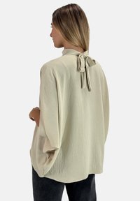 Leicht beige Bluse mit strukturierter Oberfläche, lockerem Schnitt und Schleifendetail am Halsausschnitt, kombiniert mit dunkler Hose.