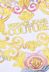 Tkanina z ozdobnymi, złotymi i różowymi wzorami barokowymi, na której wyraźnie widnieje napis "VERSACE JEANS COUTURE" w złotych literach. Białe tło.