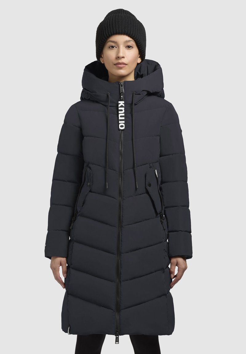 Manteau en duvet bleu marine avec capuche, comportant une fermeture éclair sur toute la longueur, deux poches extérieures et un design matelassé en chevrons ; inclut des cordons de serrage sur la capuche.