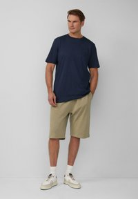 Maglietta a maniche corte blu navy abbinata a pantaloni corti beige. Il modello indossa sneakers bianche con accenti grigio chiaro e calzini bianchi alla caviglia.