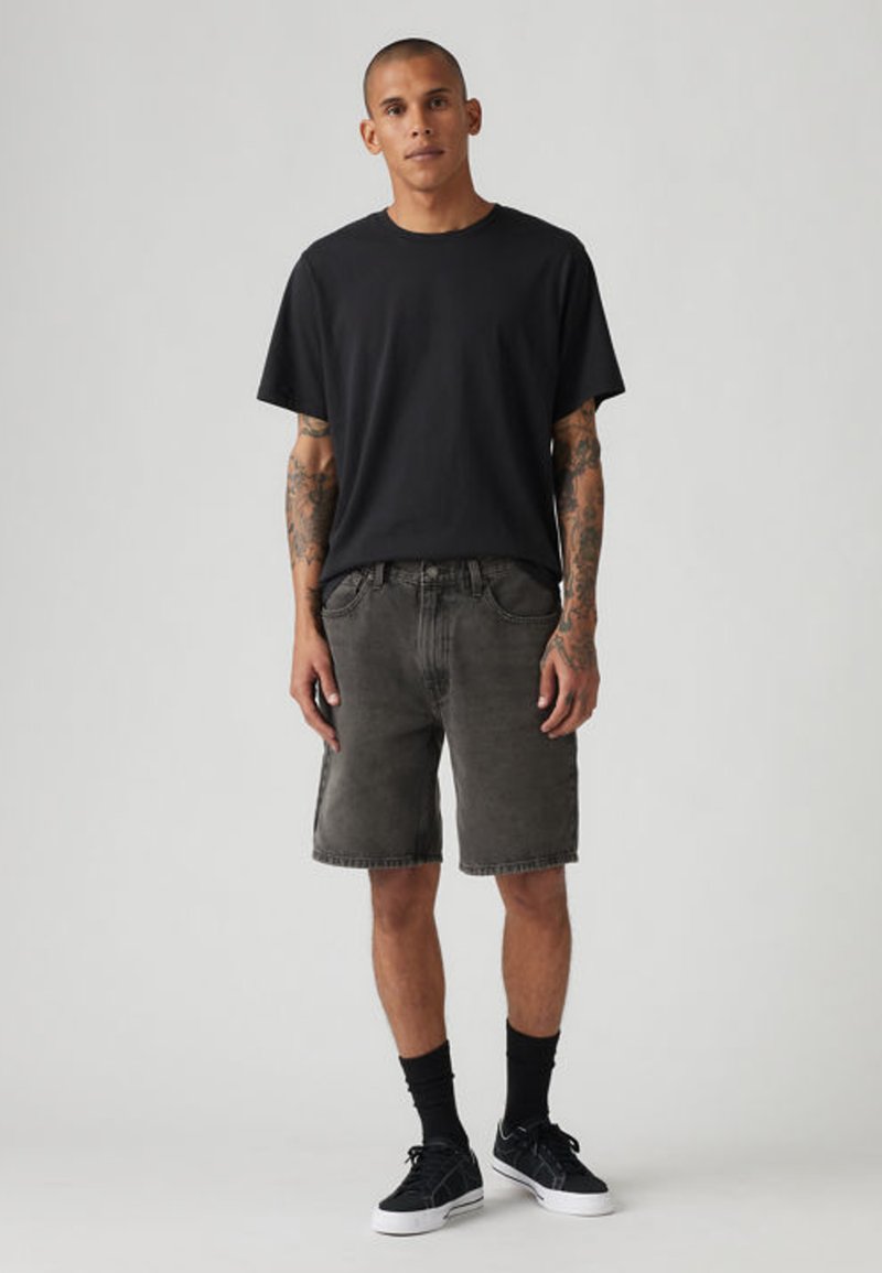 Mand med kort hår og tatoveringer iført en sort t-shirt, mørkegrå denimshorts, sorte sokker og sorte sneakers mod en grå baggrund.