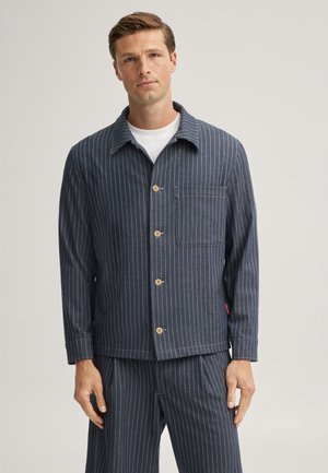 Mann trägt eine marineblaue Nadelstreifenjacke und passende Hose mit einem weißen Hemd darunter, steht vor einem schlichten Hintergrund.