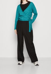 Maglione avvolgente in maglia glitter teal con maniche lunghe e dettagli di legatura, abbinato a pantaloni neri a gamba larga e scarpe sportive di colore chiaro.