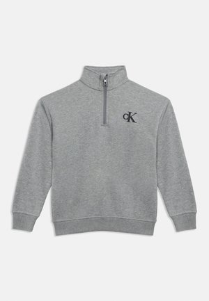 Jasnoszary sweter z długim rękawem, kołnierzem z suwakiem do ćwiartki i czarnym logo "cK" na lewej piersi.