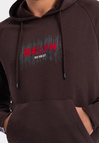 Brązowa bluza z kapturem z przednią kieszenią, z czerwonym i białym napisem "BKLYN" oraz "NEW YORK CITY." Gładka tekstura, regulowane sznurki, casualowy design.