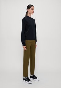 Zwarte sweatshirt met hoge hals, olijfgroene broek en zwarte sneakers met witte zolen. Glad textiel en ontspannen pasvorm.