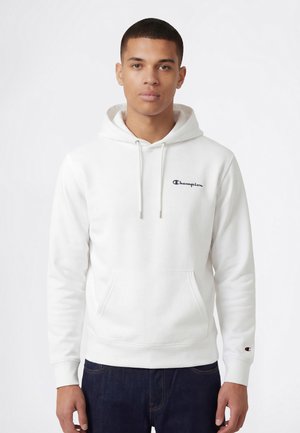 Młody mężczyzna w białej bluzie Champion z kapturem, sznurkami i kieszenią kangurką, ubrany w ciemnoniebieskie dżinsy, stojący na białym tle.