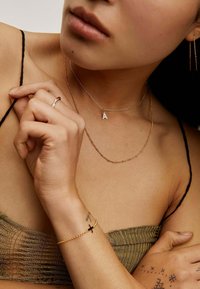 Collares en capas de oro y plata, que presentan un pequeño charm con la letra "A". Una delgada pulsera de oro con un acento de cruz. Tono de piel visible con tatuajes en los dedos.