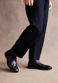Čierne kožené chelsea čižmy s elegantným dizajnom, hladkou textúrou a nízkym podpätkom, v kombinácii s tmavými rifľovými nohavicami, zobrazené v póze chôdze.