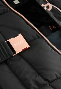 Čierna prešívaná bunda so zlatými kovovými prvkami a detailmi na zips. Interiér je podšitý mäkkou látkou, na ktorej je názov značky "BAKER."