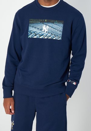 Osoba ubrana w granatową bluzę i spodnie z grafiką siedzeń stadionu oraz logo na rękawie i nogawce.