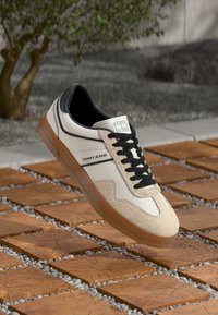 White and beige Tommy Jeans sneaker with black laces hovering over wooden square tiles outdoors.