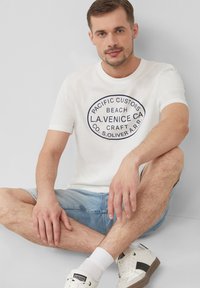 Witte katoenen t-shirt met een cirkelvormige marineblauwe grafiek met de tekst "PACIFIC CUSTOMS BEACH L.A. VENICE CA." In combinatie met lichtblauwe denim shorts.