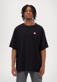 Black oversized cotton t-shirt with a round neckline and short sleeves. Features a small red logo on the left chest area.