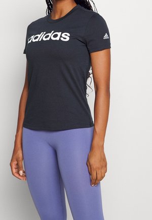 Kvinde iført en sort Adidas t-shirt med hvidt logo og lyse lilla tætsiddende leggings, stående mod en ensfarvet lys baggrund.