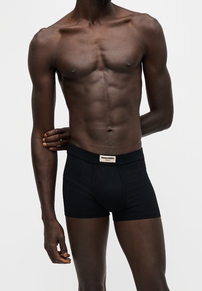 Torse masculin à la peau foncée portant un boxer noir Dsquared2 avec étiquette, sur un fond clair uni, avec les mains positionnées derrière et sur le côté.