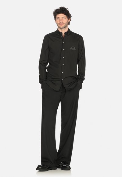 Uomo che sta in piedi con le mani in tasca, indossando un abbigliamento completamente nero che include una camicia con bottoni e logo sul petto, pantaloni larghi e scarpe nere.