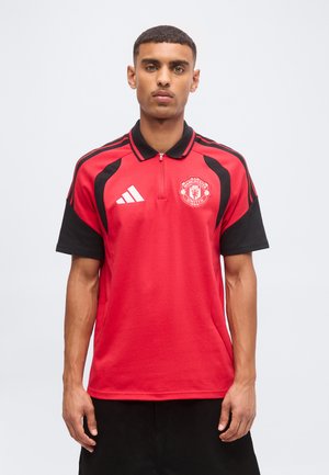 Ung mand med kort hår iført en rød og sort Manchester United sportstrøje med Adidas-logo, der står foran en ensartet lys baggrund.