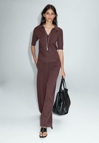 Bordeaux strikket polo-top med korte ærmer og v-hals, parret med matchende tapered bukser og en sort læder tote bag. Glat tekstur, minimalistisk design.